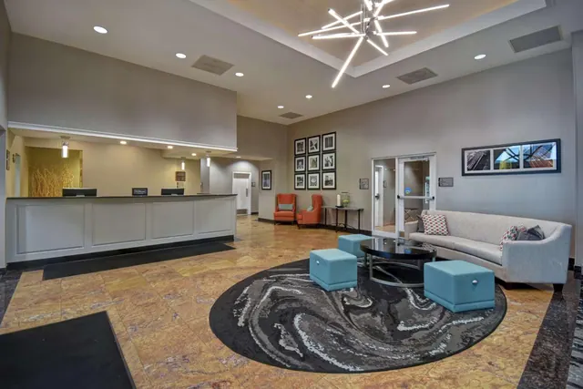 A hotel lobby with a reception desk on the left, a grey sofa, orange armchairs, teal ottomans and a round patterned rug under a modern chandelier.