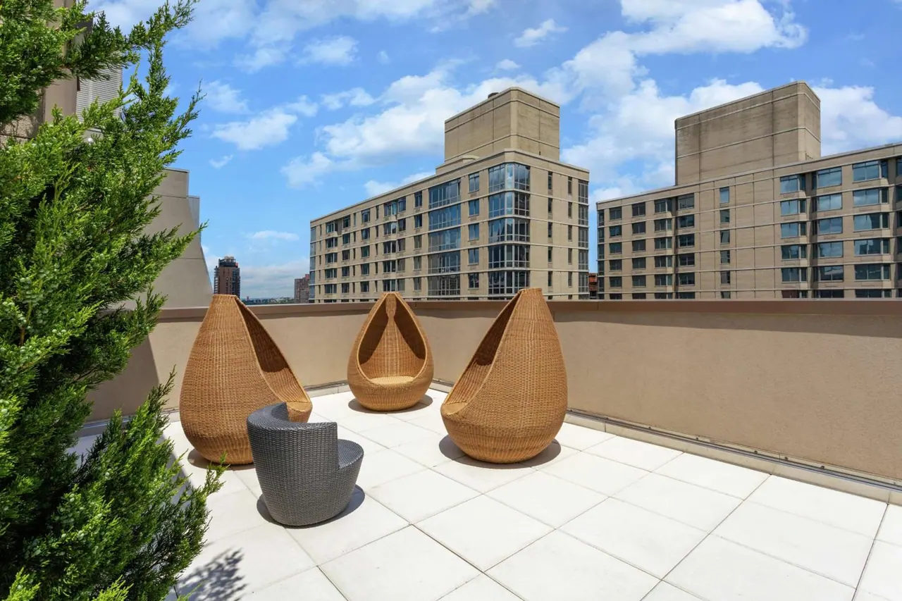 A rooftop terrace with light tiled flooring, three woven pod chairs and a grey wicker chair, with apartment buildings visible beyond the low wall.
