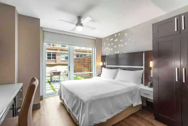 A bedroom with a made king bed, a desk and chair, a wardrobe, and glass doors opening onto a small courtyard patio with a brick building behind.