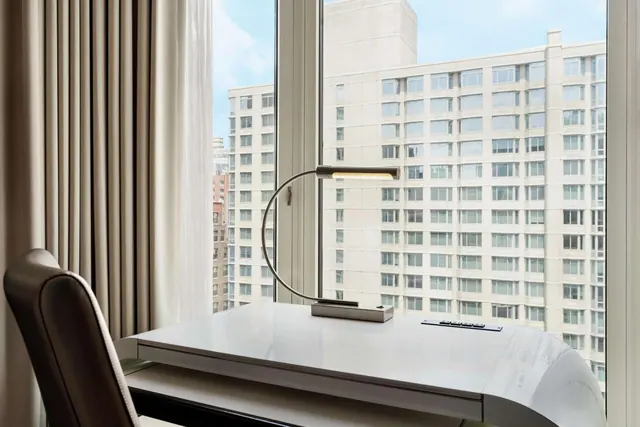 A white curved desk with a desk lamp and a dark leather chair stands in front of a window overlooking a tall residential building.