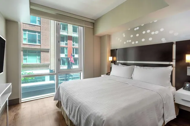 A bedroom with a king bed, a wall-mounted television and floor-to-ceiling windows looking onto a brick apartment building across the street.