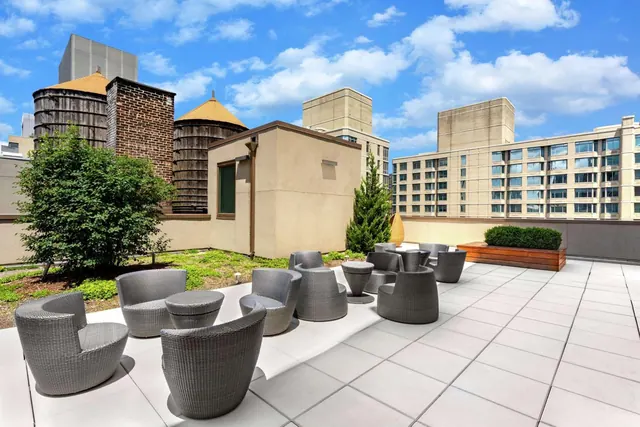 A rooftop terrace with grey wicker armchairs and low tables, with surrounding city buildings and water towers visible under a blue sky.
