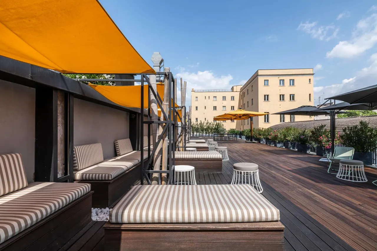 A rooftop terrace with striped daybeds, yellow shade sails and potted plants, with neighbouring buildings visible beyond.