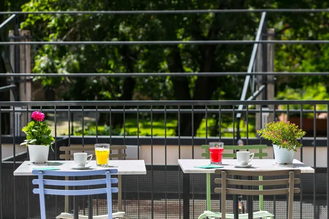 Two small marble-topped tables with coloured metal chairs, drinks and potted flowers sit on a balcony with a metal railing overlooking trees and a lawn.