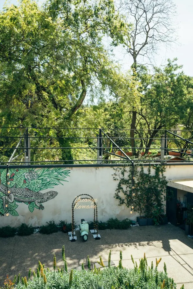 A paved courtyard has a painted lizard mural on the wall, a mint scooter beside a light arch sign, and tall trees behind a metal railing.