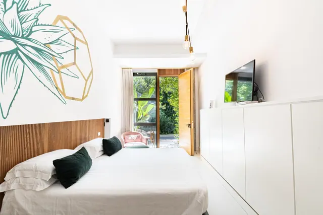 A bedroom with a made double bed, a wall-mounted television, white storage cabinets, a pink armchair, and a glass door opening onto a garden.