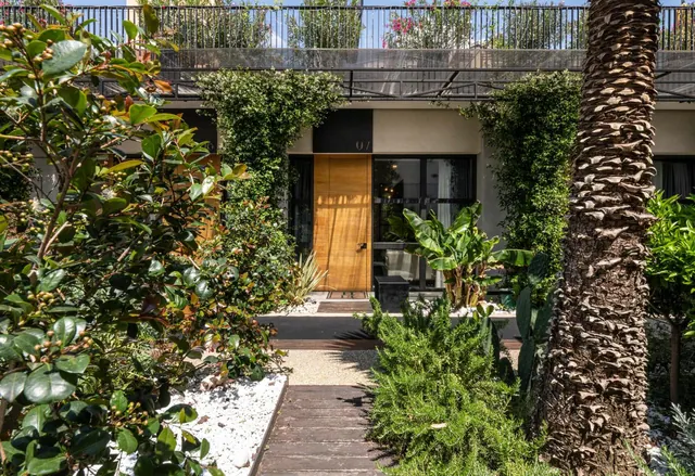 A wooden pathway through a planted garden leads to a wooden door marked 07 beneath an upper balcony with a black railing, with a palm trunk in the foreground.