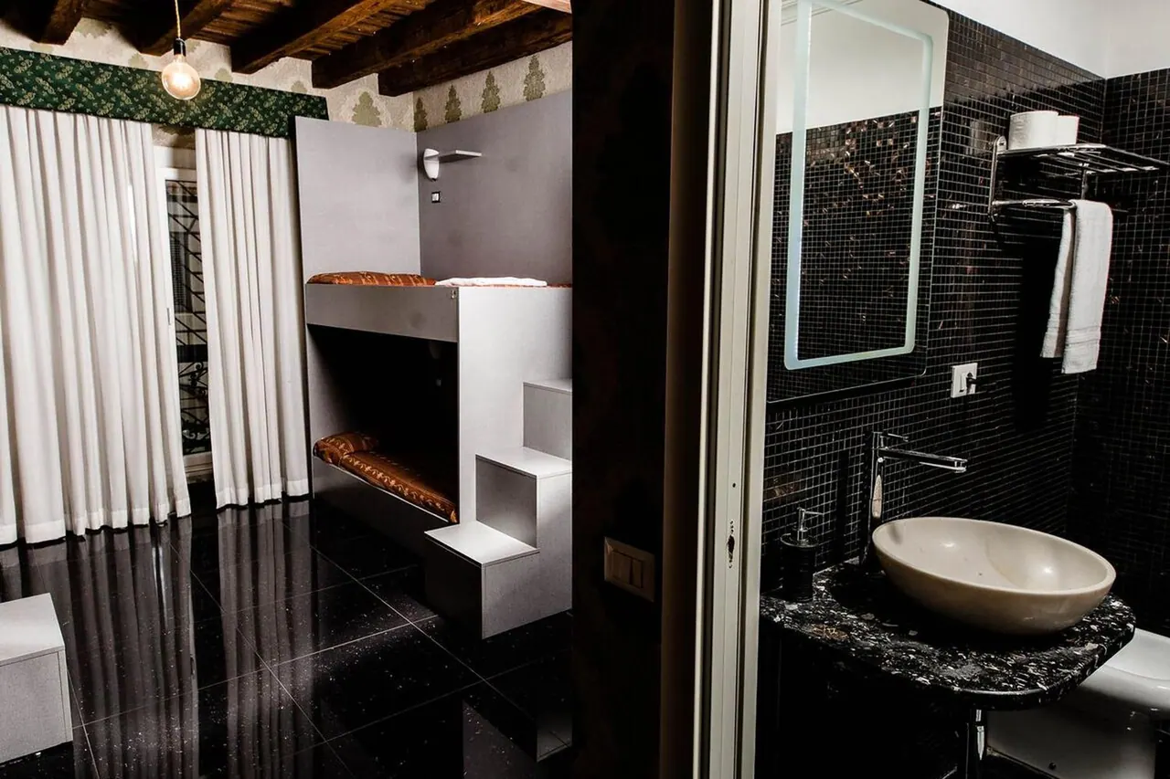 A bedroom with a built-in bunk bed and white curtains sits beside a bathroom with black mosaic tiles, a round basin and a backlit mirror.