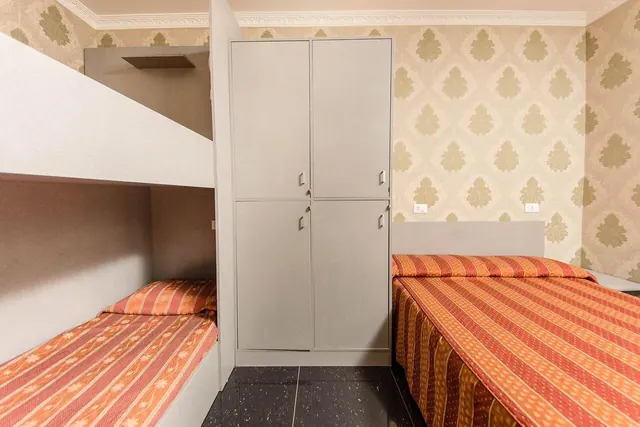 A bedroom with a bunk bed on the left, a tall grey wardrobe in the centre and a bed with orange striped covers on the right, against damask wallpaper.