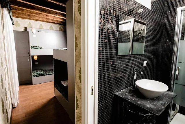 A black mosaic-tiled bathroom with a vessel sink on a dark marble vanity, a glass shower enclosure on the right, and a bunk-bed room visible through the open doorway.
