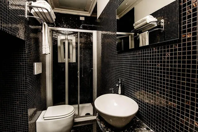 A bathroom with black mosaic tiles, a glass-door shower enclosure, a wall-mounted toilet, a vessel sink on a dark marble counter, and heated towel rails.