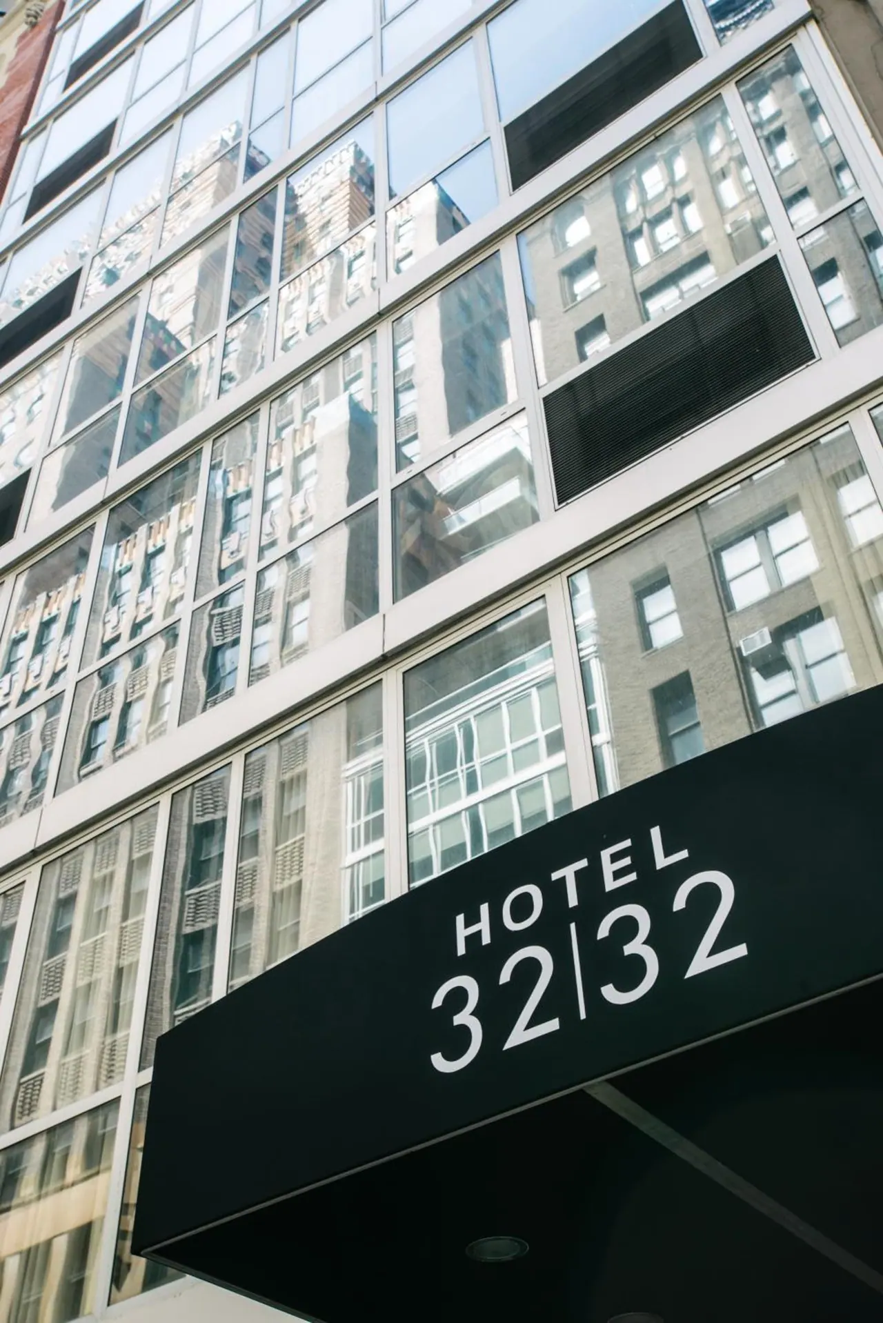 Exterior of Hotel 32|32 showing a glass facade that reflects neighbouring buildings, with a black entrance awning bearing the hotel name.
