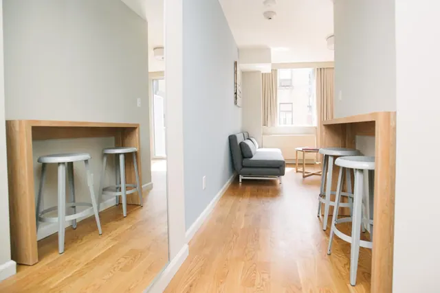 A narrow room with wood floors, two wooden counters with grey stools, a grey sofa, and a window with curtains.