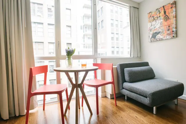 A small seating area with two red chairs and a round wooden table holding a flower vase, beside a grey sofa chair and a large window overlooking neighbouring buildings.