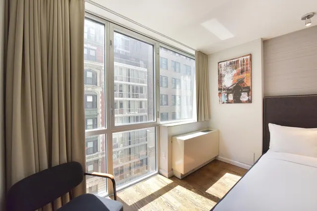 A hotel bedroom with a bed, a chair beside floor-to-ceiling windows looking onto urban buildings, and sunlight falling on the wooden floor.