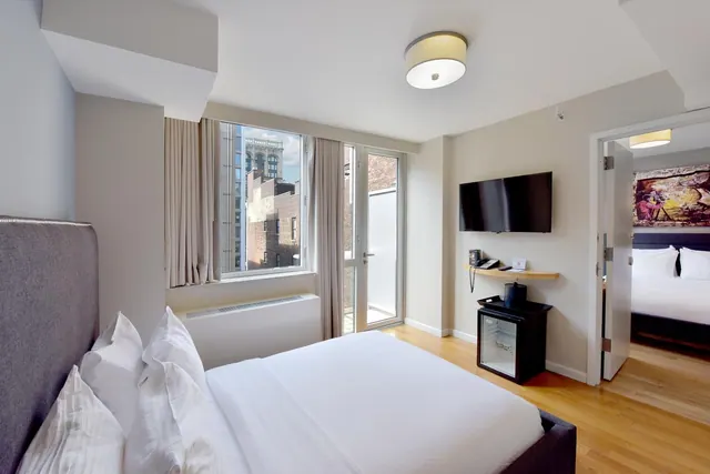 A bedroom with a made bed, a wall-mounted television, a minibar fridge, and a window and glass door looking onto brick buildings across the street.