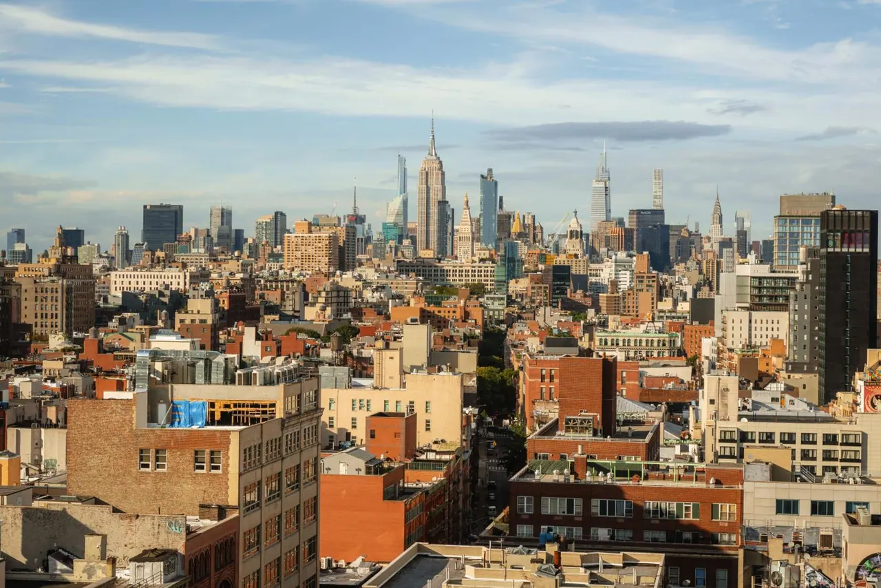Elevated view over dense brick and stone buildings toward a Manhattan skyline with the Empire State Building visible in the distance.