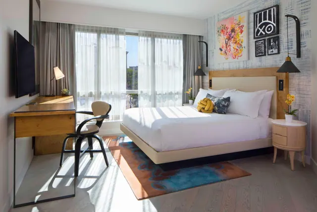 A bedroom with a king bed, a wall-mounted television, a wooden desk and chair, and floor-to-ceiling windows with sheer curtains looking onto a street with trees.
