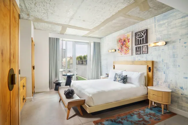 A bedroom with a made king bed, a wooden wardrobe, and glass doors opening onto a small outdoor terrace with a round table and stool.