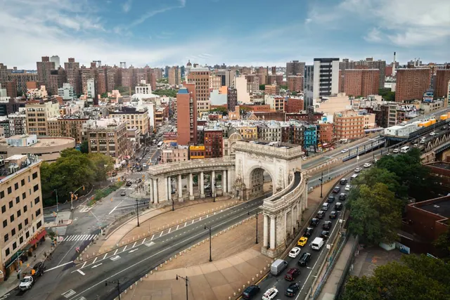 Aerial view of a white stone arch and colonnade beside a wide avenue with traffic, with dense apartment buildings stretching into the distance under a partly cloudy sky.