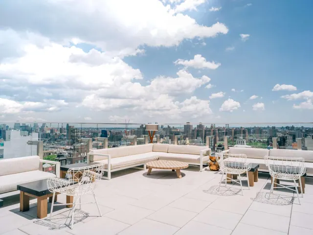A rooftop terrace with white sofas, wire chairs and a low wooden table overlooks a city skyline under a partly cloudy sky.