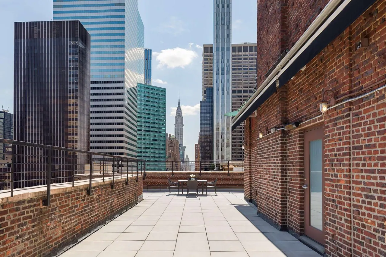 A brick-walled rooftop terrace with a small table and chairs overlooks high-rise buildings, with the Chrysler Building visible in the distance.