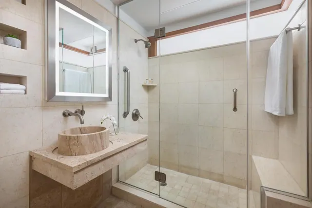 A bathroom with a walk-in glass shower, a stone basin on a floating vanity, and a backlit mirror.