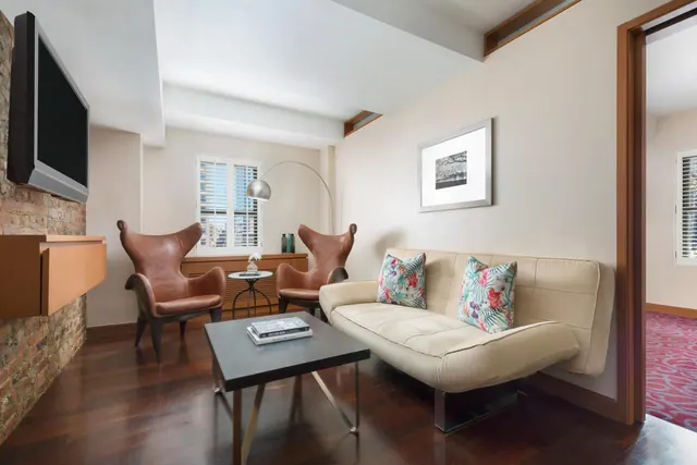 A seating area in a hotel suite with a wall-mounted television, two leather armchairs, a cream sofa, and a coffee table.