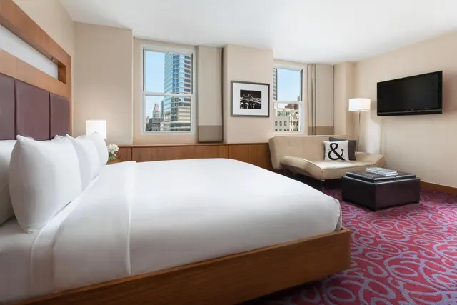 A hotel bedroom with a king bed, a cream sofa, a wall-mounted television, and windows showing city high-rises.