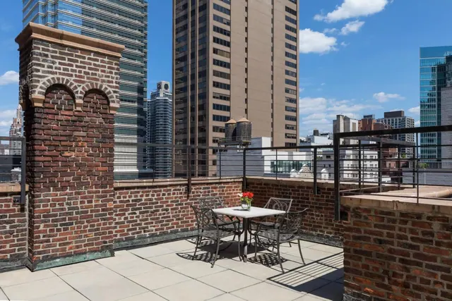A brick-walled rooftop terrace with a metal bistro table, four chairs and a vase of red flowers overlooks high-rise buildings under a blue sky.