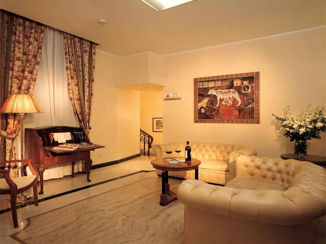 A seating area with cream leather armchairs and a sofa, a round wooden table holding two wine glasses and a bottle, a writing desk by the curtained window, and a framed painting on the wall.