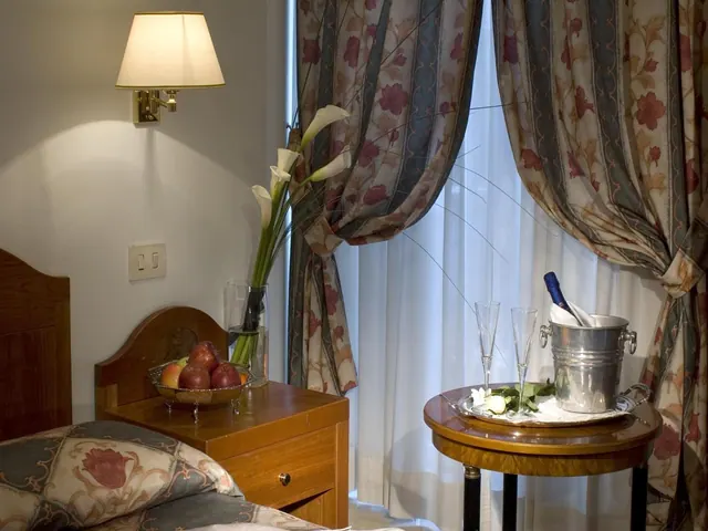 A bedroom with a wall lamp, a bed beside a nightstand holding a fruit bowl and calla lilies, and a round side table with a champagne bucket and two flutes in front of sheer-curtained windows.