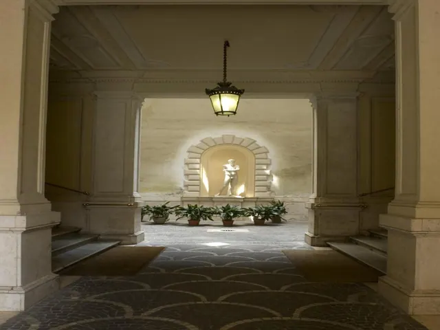A columned entrance passage with a hanging lantern, potted plants, a lit statue in a niche and a patterned stone floor.