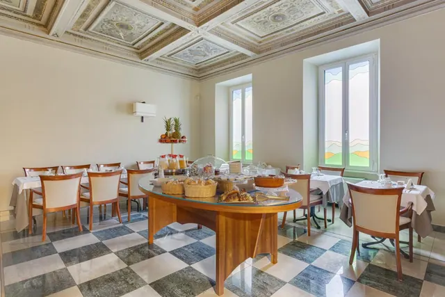 A breakfast dining room with a painted coffered ceiling, checkered floor, laid tables and a central buffet table with bread, pastries and fruit.