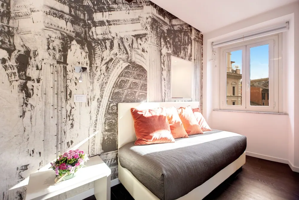 A bedroom with a double bed topped with coral cushions, a black-and-white mural of a Roman arch on the wall, a white side table with flowers, and a window looking onto neighbouring buildings.