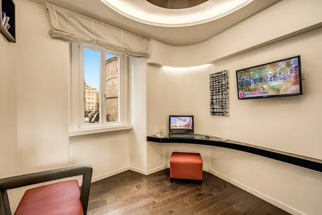 A small room with a window onto nearby buildings, a curved wall desk with a laptop, a wall-mounted television and red upholstered seating.