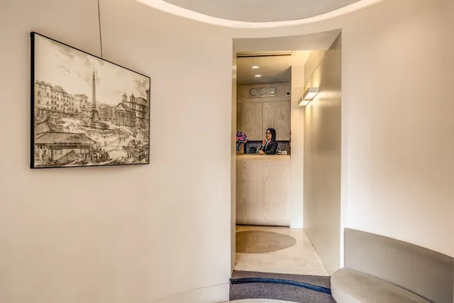 A curved hallway leads to a reception desk where one staff member sits behind the counter, and a framed engraving of a piazza with an obelisk and a domed building hangs on the left wall.