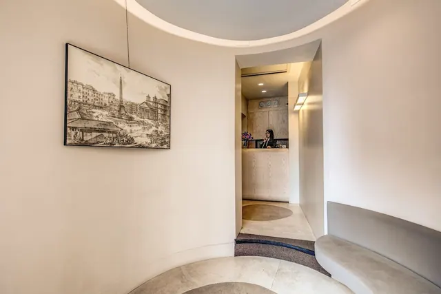 A curved entrance area with a framed engraving of a Roman piazza on the wall, a curved bench on the right, and a receptionist behind a wooden desk at the far end.