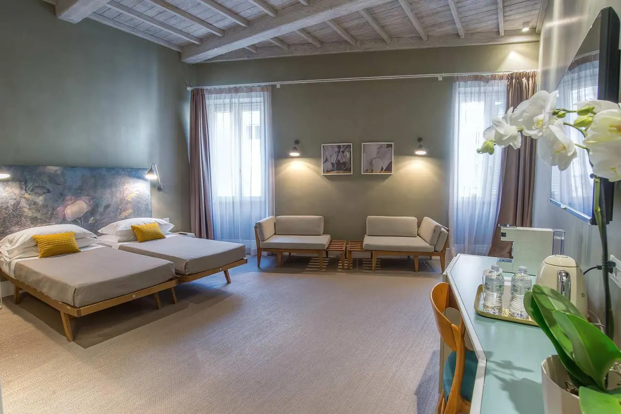 A bedroom with twin beds and yellow cushions, armchairs by the windows, a wall-mounted television and a desk with a kettle and bottled water.