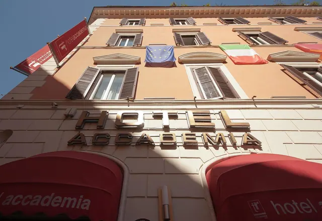 Low-angle view of the hotel facade showing the Hotel Accademia sign, red awnings, red banners, and EU and Italian flags on the upper floors.