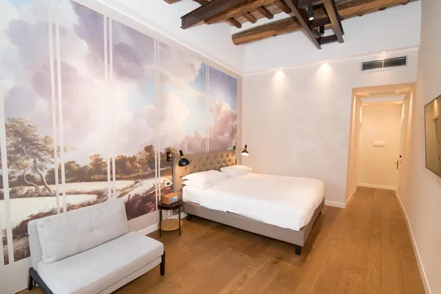 A bedroom with a double bed, a painted landscape mural on one wall, a white armchair and an exposed wooden beamed ceiling.