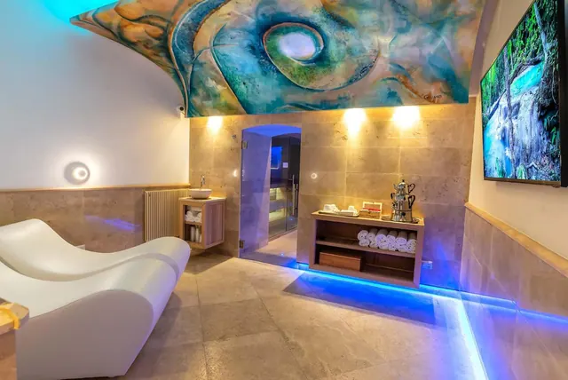A spa room with a painted vaulted ceiling, white loungers, a wall-mounted television showing a forest waterfall, and a shelf of rolled towels.