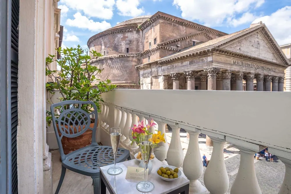 A small balcony with a metal chair, potted plant and a side table holding champagne flutes, flowers and olives overlooks the Pantheon.