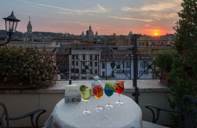 A round table with a white cloth holds four cocktail glasses and a small box on a rooftop terrace overlooking the city at sunset.