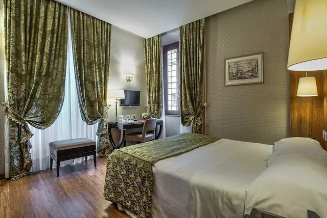 A hotel bedroom with a double bed, green damask curtains, a writing desk with a television, and a framed print on the wall.