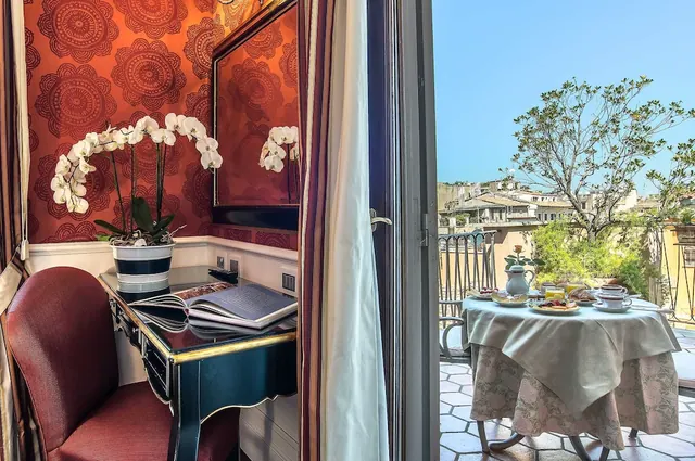 A desk with an open book and a white orchid sits beside a door opening onto a balcony with a breakfast table and rooftops beyond.