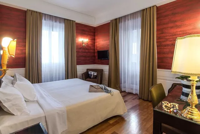 A bedroom with a double bed, red textured walls, a wall-mounted television, an armchair and a desk with a table lamp.