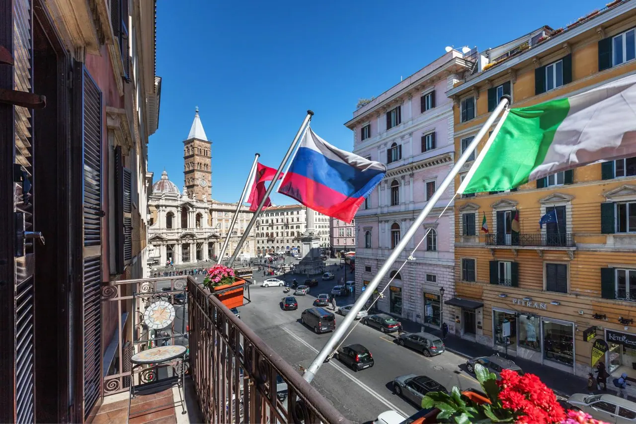 View from a balcony over a street and square with parked and moving cars, the bell tower of Santa Maria Maggiore, and Russian and Italian flags on poles.
