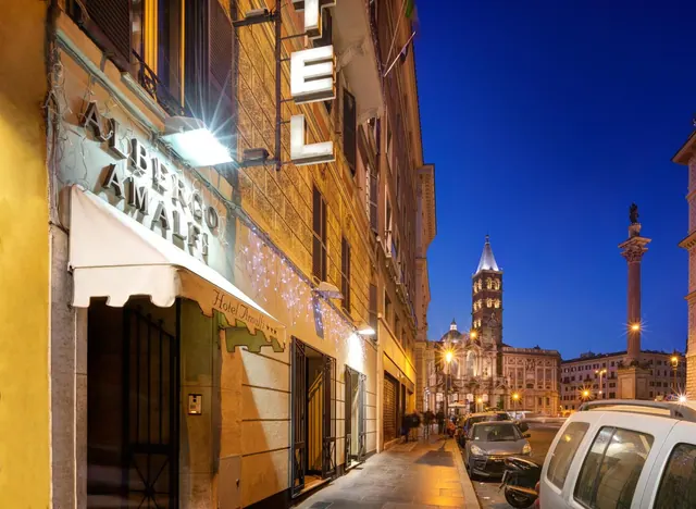 Evening street view of the Albergo Amalfi hotel facade in Rome, with its illuminated sign and the bell tower of Santa Maria Maggiore visible beyond.
