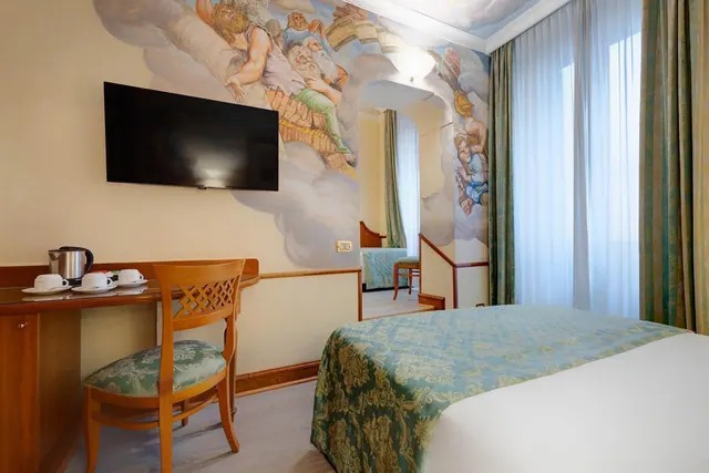 A hotel bedroom with a bed under a teal damask cover, a wall-mounted television, a wooden desk with a kettle and cups, and a painted fresco covering the wall and ceiling.